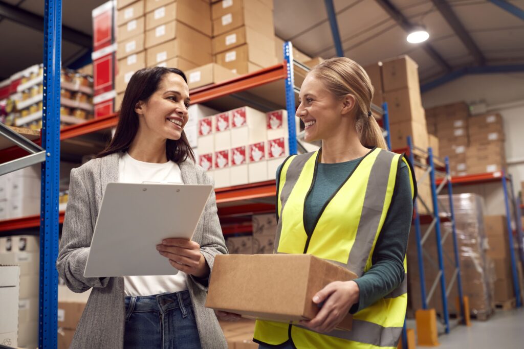 Zwei Frauen in einem Lagerraum, die ein Paket halten und eine Checkliste durchsehen.