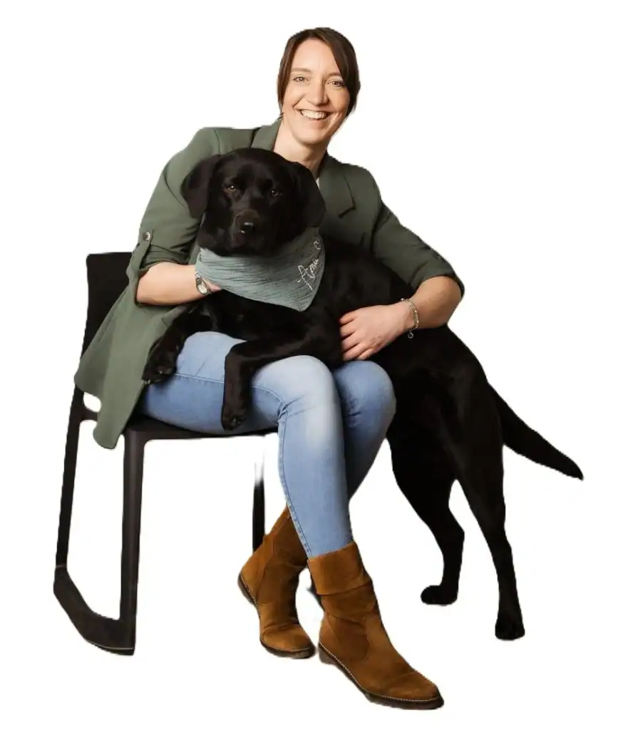 Porträt von Maria Röming, sitzend auf einem Stuhl. Ihre schwarze Labradorhündin Amie trägt ein grünes Halstuch mit ihrem Namen und hat die Vorderpfoten auf Marias Schoß.