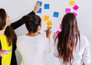 Drei Frauen stehen vor einem Whiteboard und sammeln Ideen mit Haftnotizen.